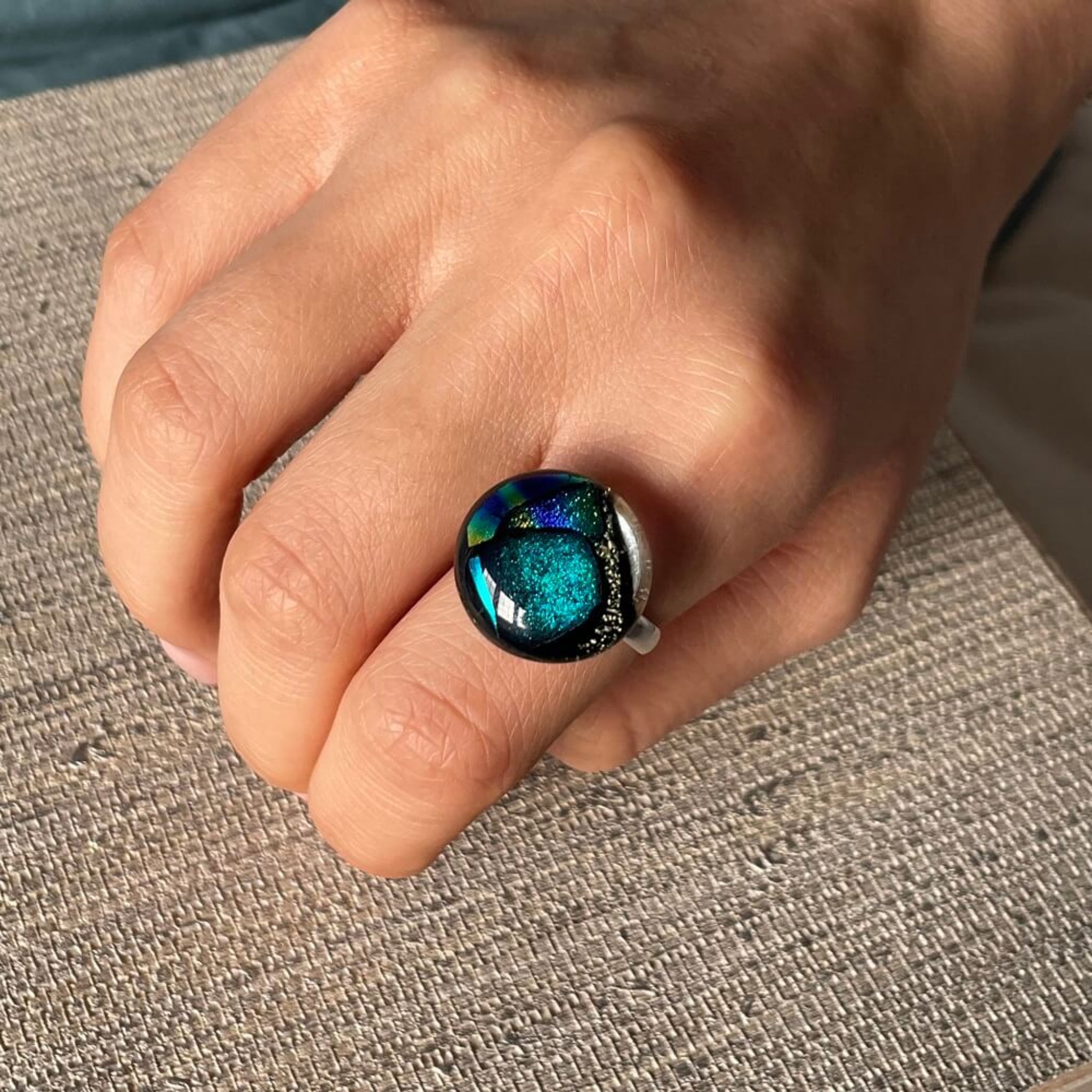 woman wearing a ring, Close-up of a round jewel statement ring, colourful dichroic fused glass ring, yellow, blue, glittering pink glass, with purple orchids in the background, on adjustable stainless steel ring base