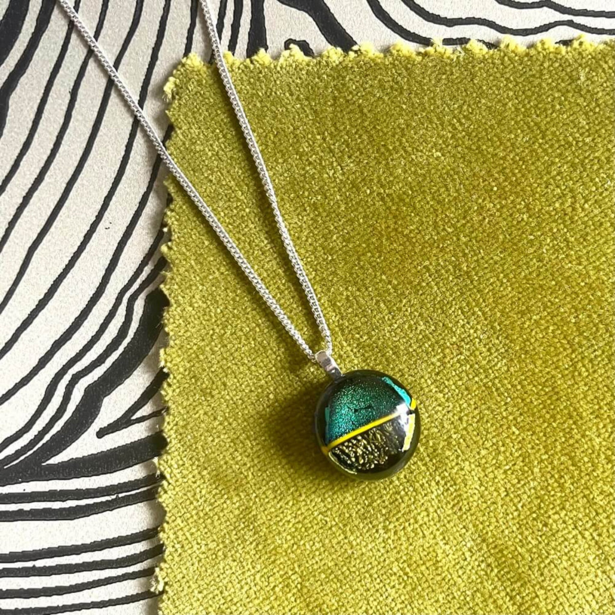 green and gold  glittering dichroic fused glass pendant on sterling silver chain on green velvet, with black patterned background