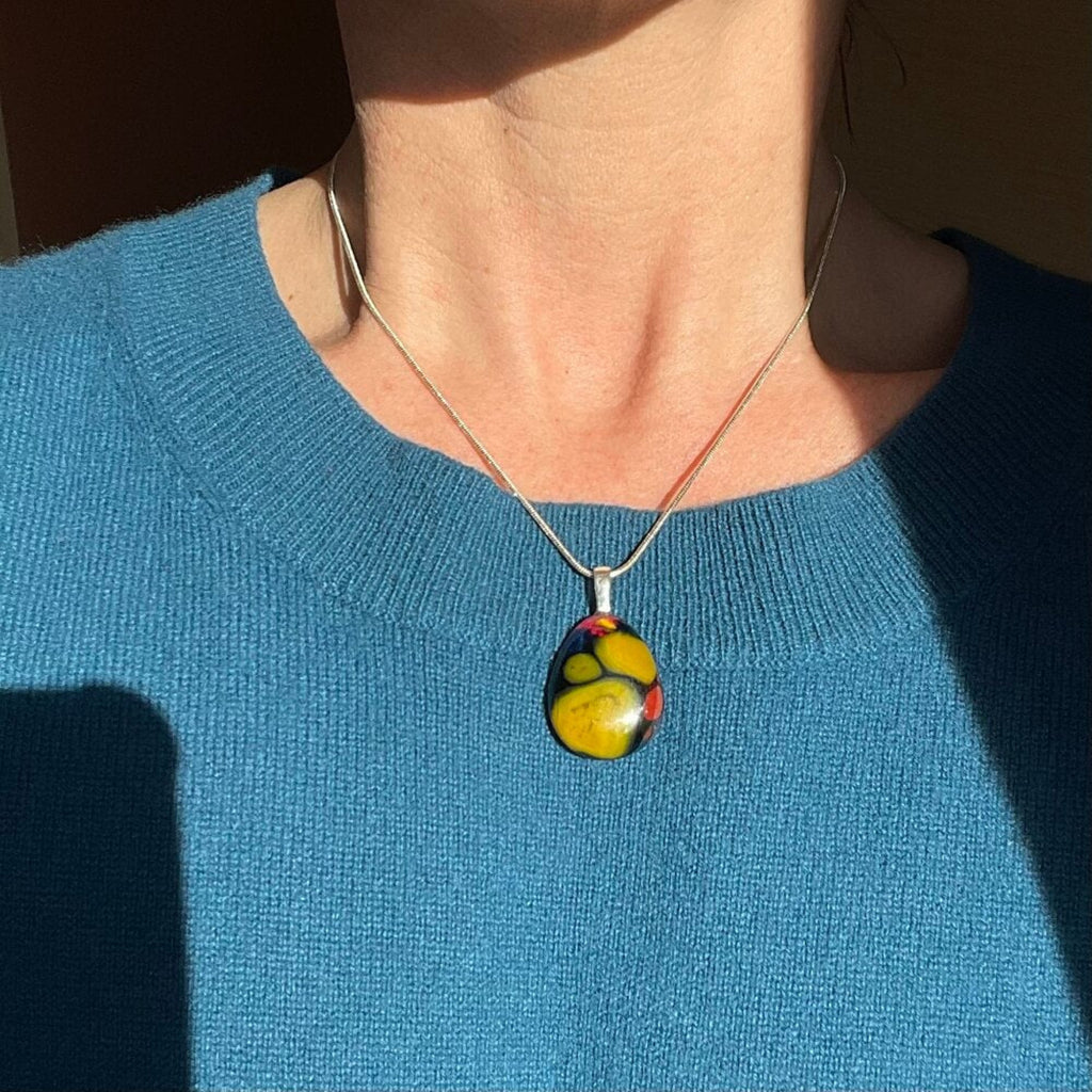 Person wearing a colorful fused glass art pendant, yellow, blue, pink fused glass, with sterling silver chain and bail, necklace against a blue sweater.